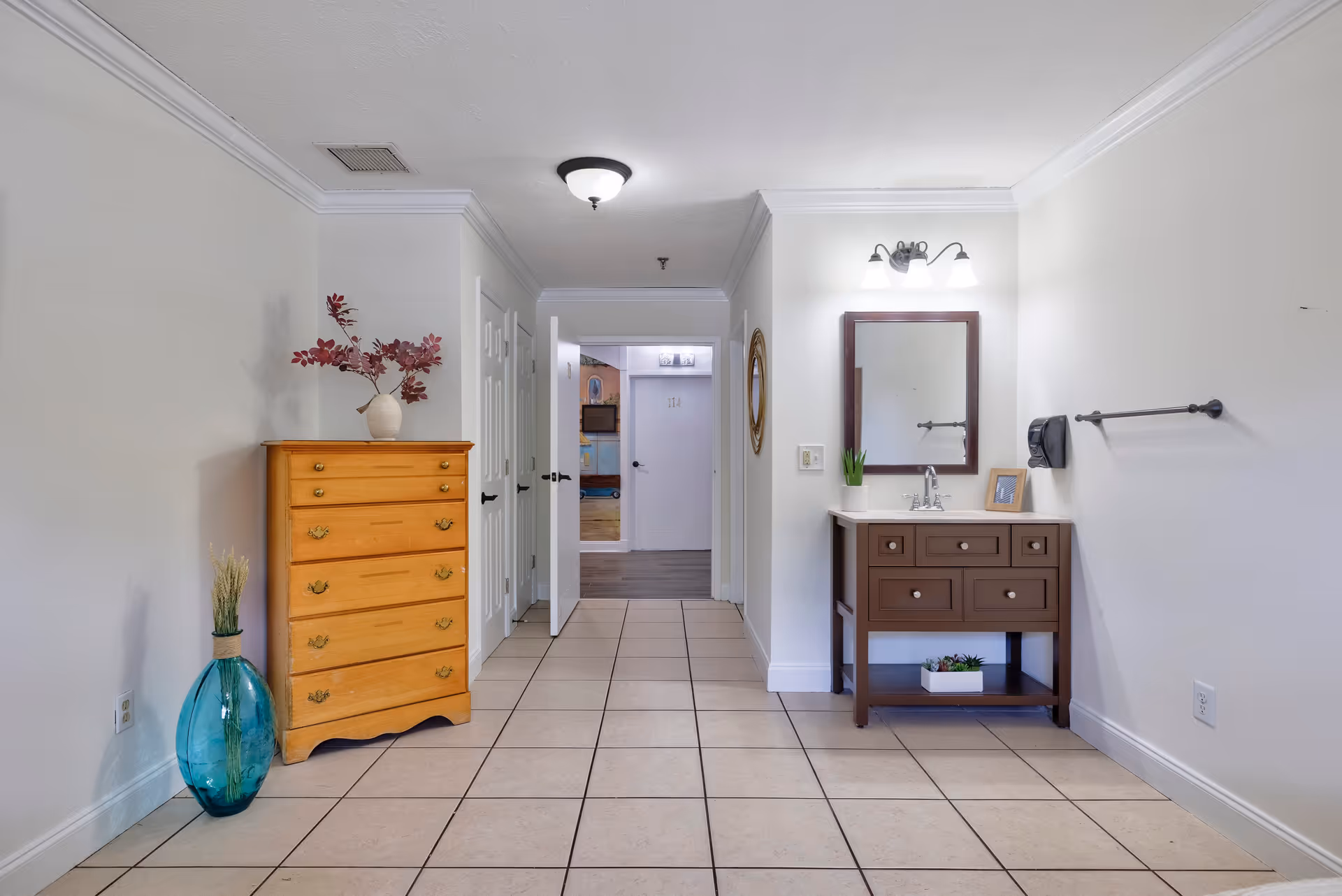A hallway area in a senior living facility with beige tiled floor and white walls. On the left side, there is a wooden chest of drawers with a vase holding red leaves on top and a large blue glass vase with dried plants beside it. On the right side, there is a brown vanity with a white countertop, a mirror above it, and a three-light fixture. A towel rack and a soap dispenser are mounted on the wall next to the vanity. In the background, an open door reveals another room with a door numbered 114.