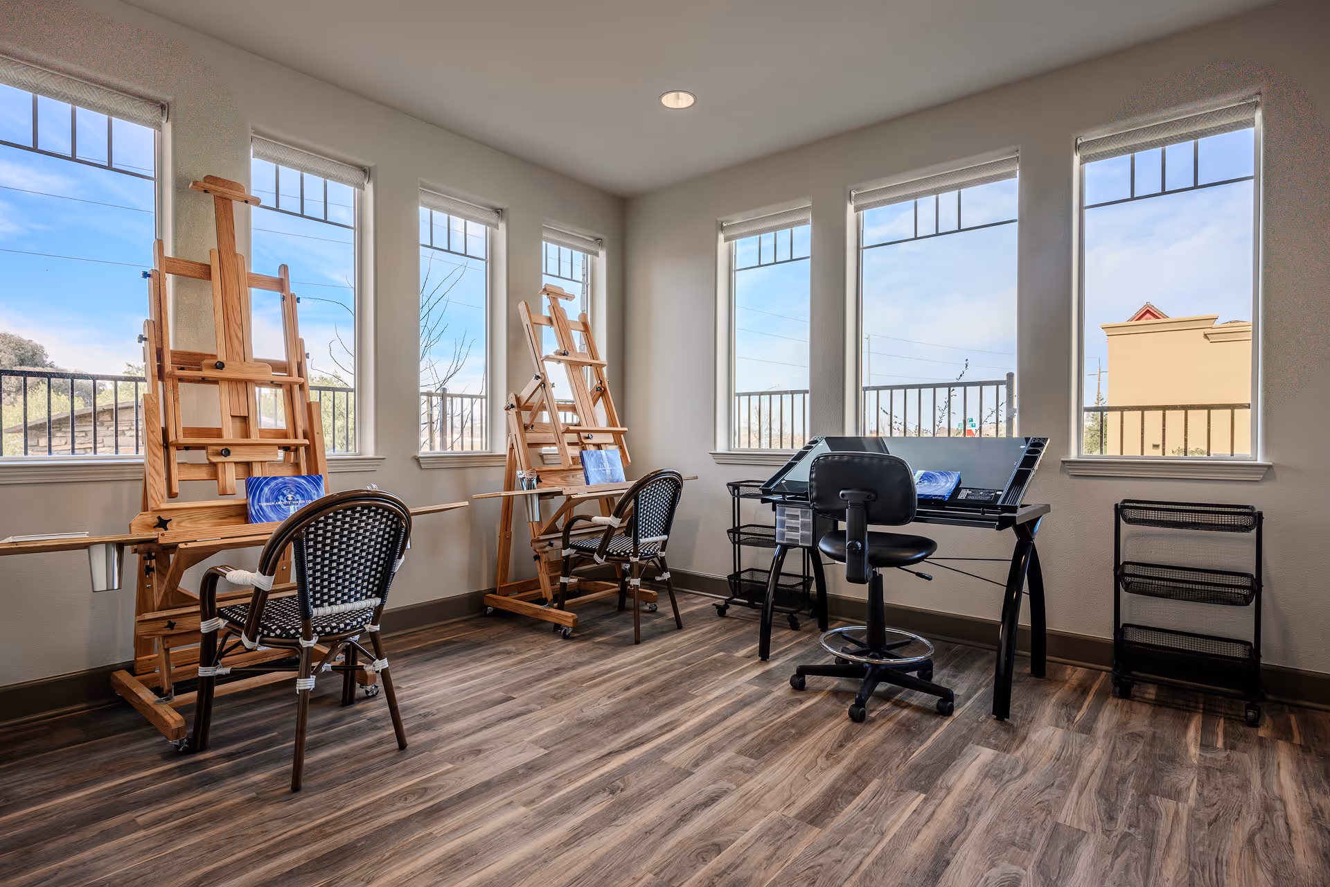 A bright art studio room with large windows letting in natural light. The room features two wooden easels with chairs in front of them, and a black drafting table with a black swivel chair. The floor is wood-patterned, and the walls are light-colored.