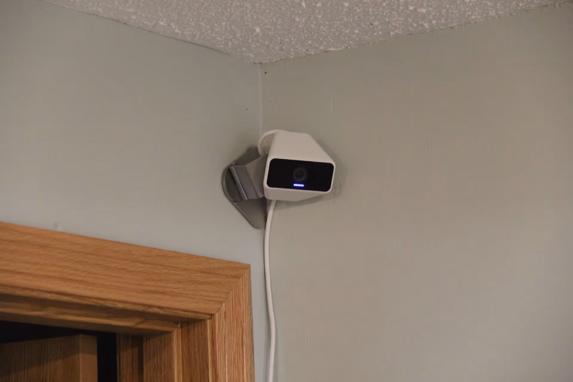 A white security camera mounted on a gray wall in the corner above a wooden door frame, with a visible white power cable hanging down.
