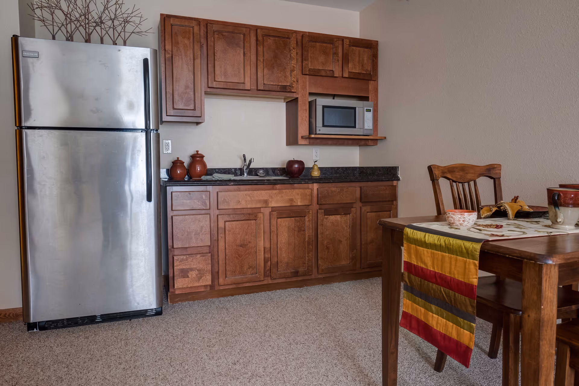 A small kitchen area with wooden cabinets, a stainless steel refrigerator, a microwave, and a countertop with a sink. To the right, there is a wooden dining table with a colorful striped table runner and some cups and bowls on top.