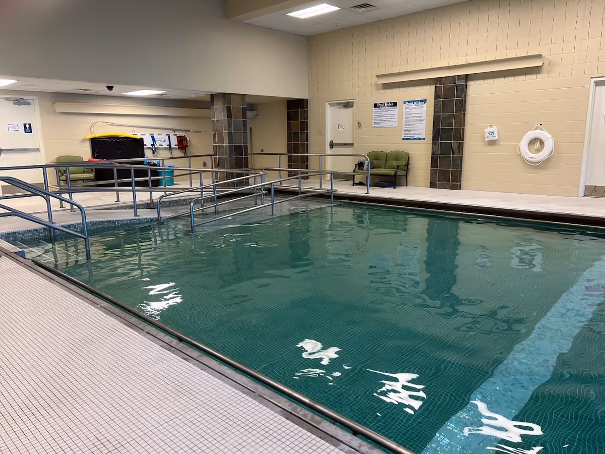 Indoor swimming pool with metal handrails, tiled deck, and a seating area in a community facility.