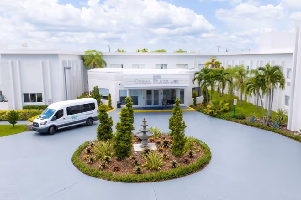 Front exterior view of Coral Plaza facility showing a circular driveway with a landscaped island featuring a fountain and various plants. A white shuttle van is parked near the entrance, and palm trees and other greenery surround the building under a partly cloudy sky.