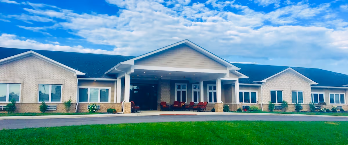 Front exterior view of a single-story assisted living facility building with a covered entrance, multiple windows, a paved driveway, and a well-maintained green lawn under a partly cloudy sky.
