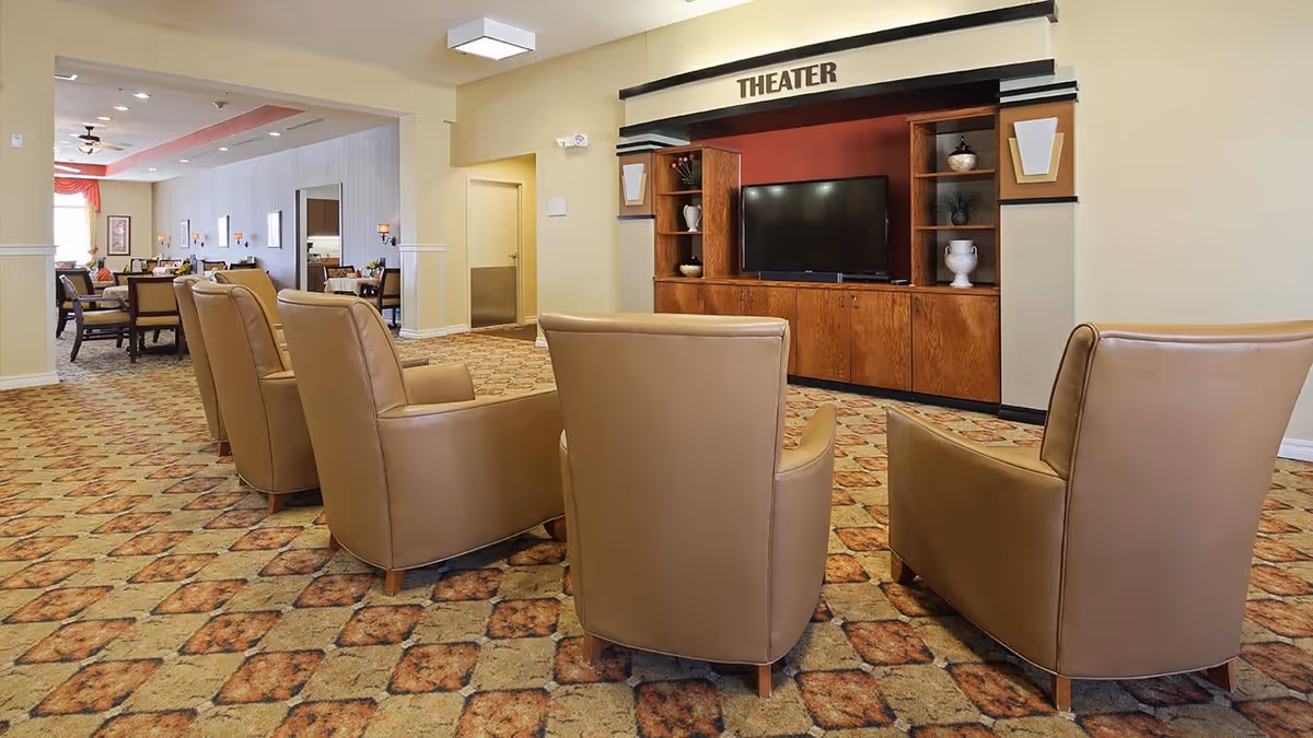A cozy theater room in an assisted living facility featuring four tan leather armchairs arranged in front of a flat-screen TV mounted on a wooden entertainment center labeled 'THEATER'. The room has patterned carpet flooring and an adjacent dining area visible in the background.