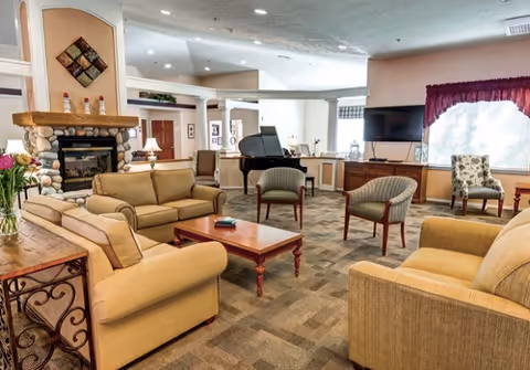 A spacious living room area in a senior living facility with beige sofas and armchairs arranged around a wooden coffee table. There is a stone fireplace with decorative items on the mantel, a grand piano in the background, a flat-screen TV mounted on the wall, and large windows with maroon curtains letting in natural light.