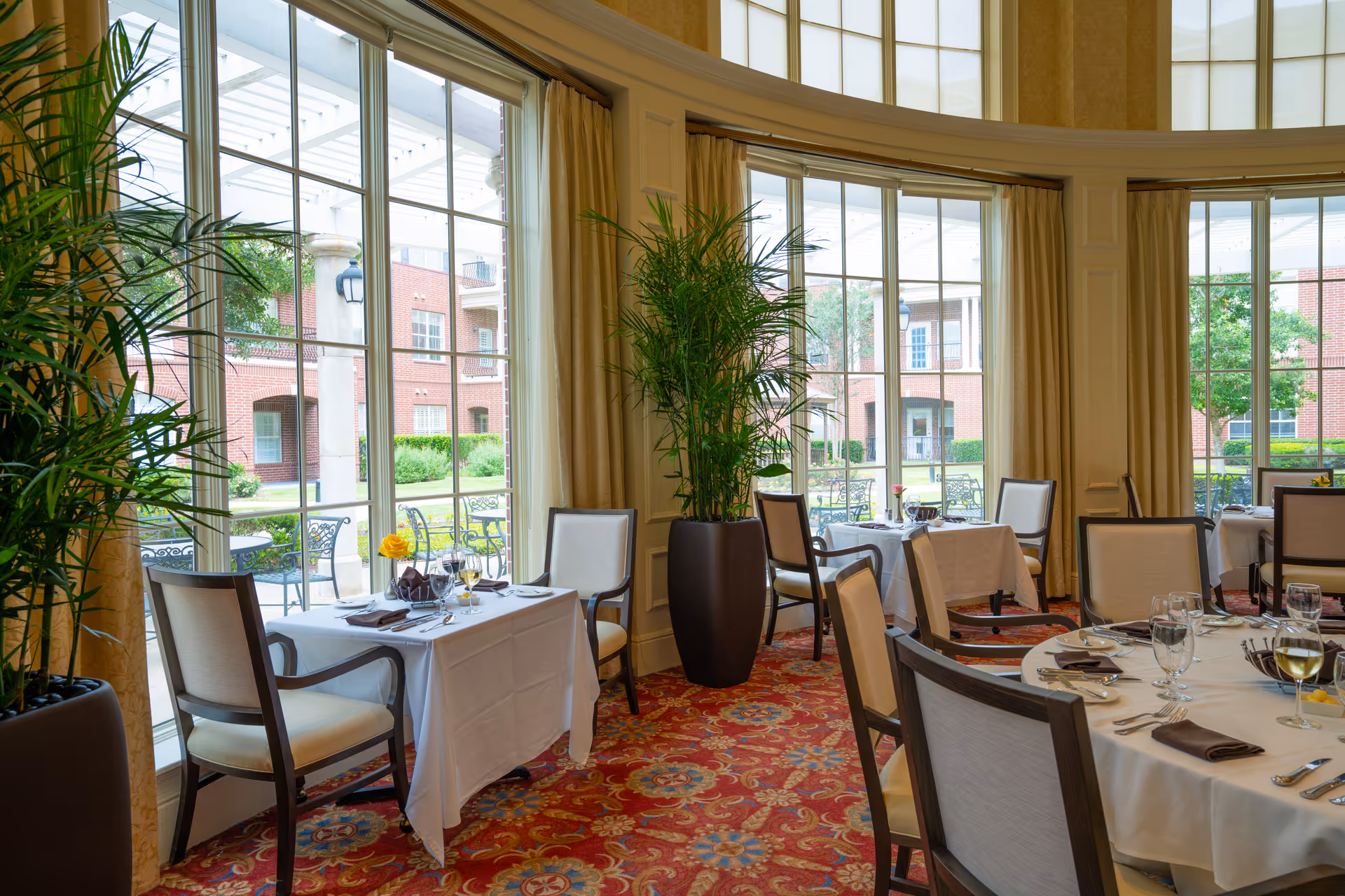 A bright dining room with large floor-to-ceiling windows showing an outdoor patio area. The room features tables covered with white tablecloths, set with glasses, plates, and napkins. There are cushioned chairs around the tables and tall potted plants near the windows. The carpet has a red and gold intricate pattern.
