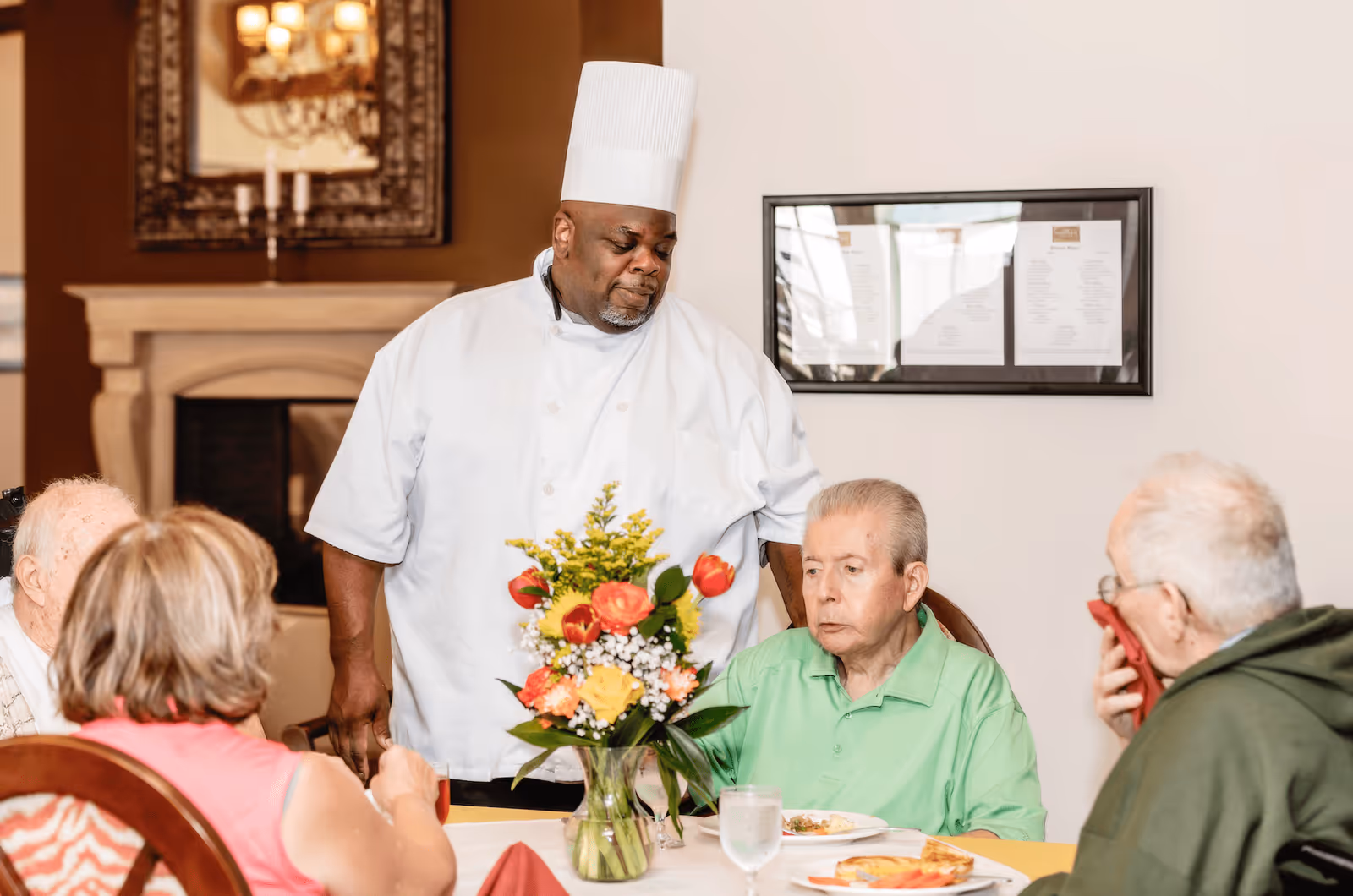 A chef in a white uniform and tall chef hat stands beside a dining table where three elderly people are seated. The table has a vase with colorful flowers, plates with food, and glasses of water. The setting appears to be a dining room with a fireplace and a framed menu on the wall in the background.