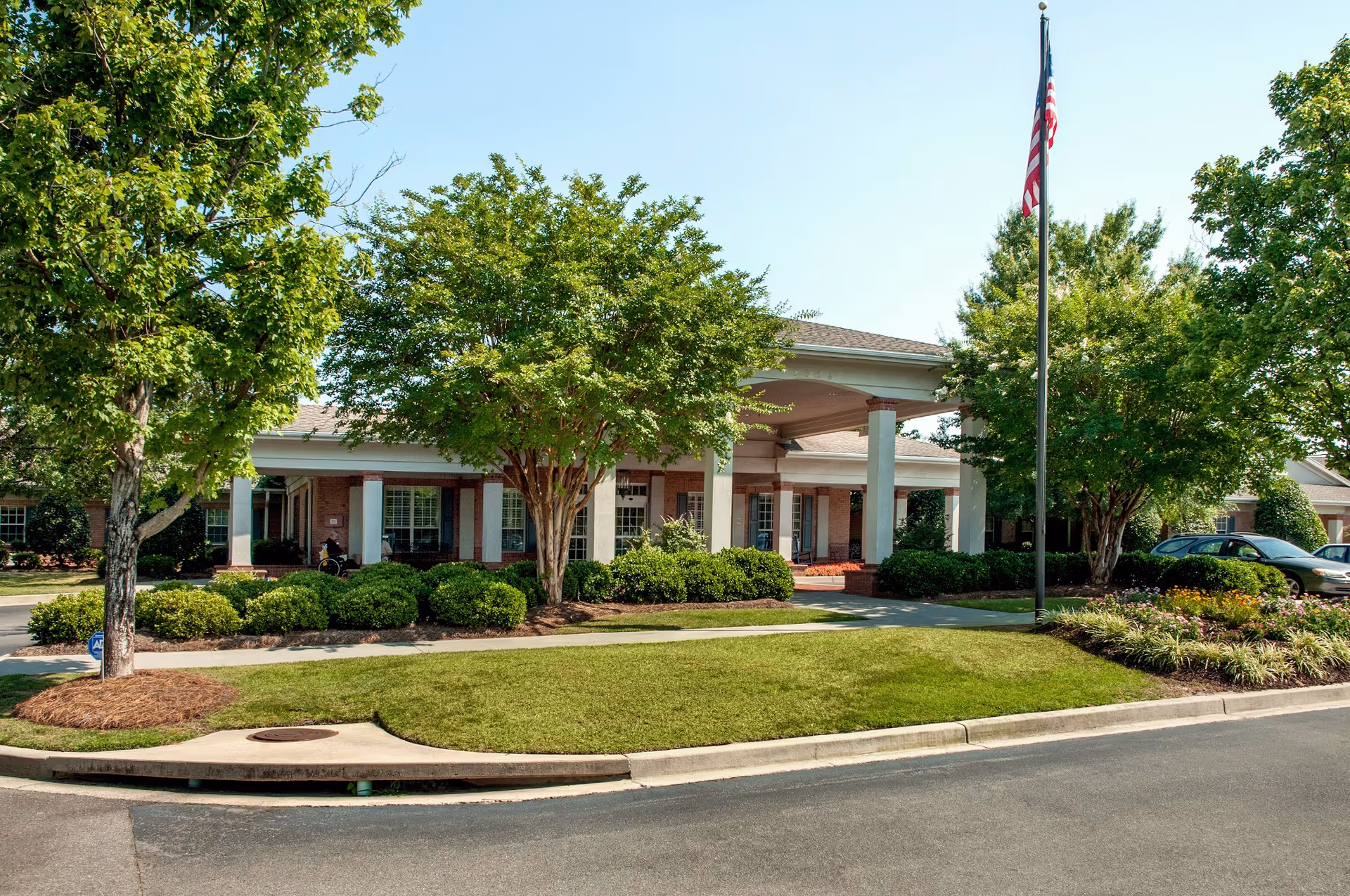 Front exterior view of Life Care Center of Columbia, South Carolina, showing a single-story brick building with white columns, surrounded by green trees and bushes, a well-maintained lawn, and an American flag on a flagpole.