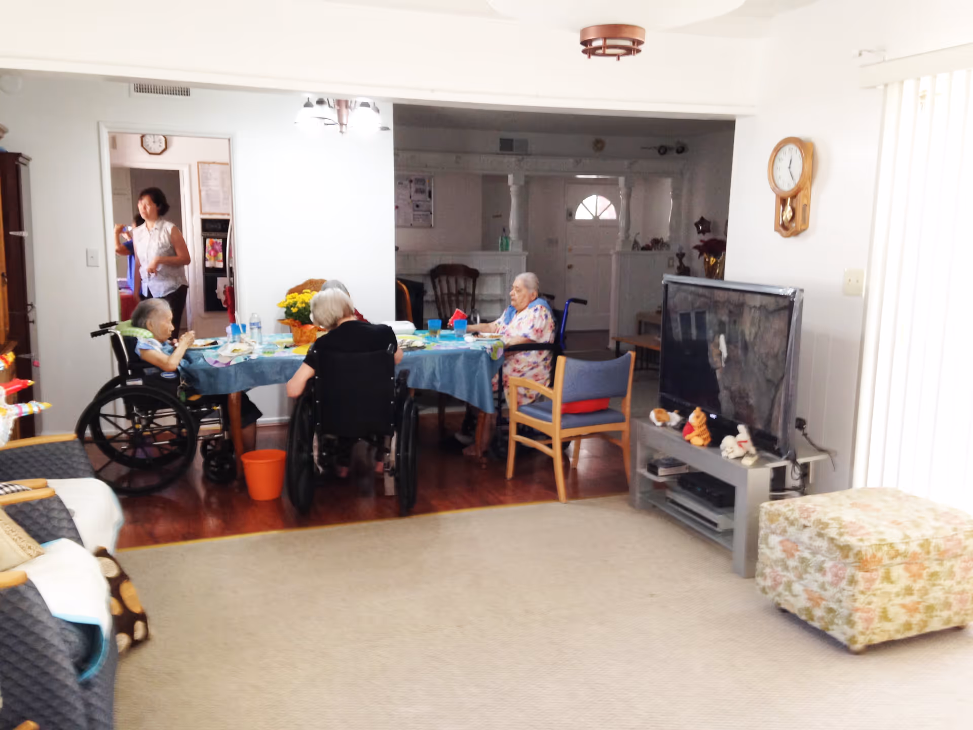 A group of elderly people, some in wheelchairs, sitting around a dining table covered with a blue tablecloth in a residential care facility. A caregiver stands nearby in the background. The room has wooden flooring, a television on a stand with stuffed animals, a floral ottoman, and a wall clock.