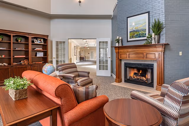 A cozy living room area in a senior living facility featuring a red sofa, two striped armchairs, a wooden coffee table, and a wooden side table with a potted plant. There is a fireplace with a wooden mantel and a painting above it. A bookshelf with various items and a globe is visible on the left side. Double glass doors lead to a bright hallway in the background.
