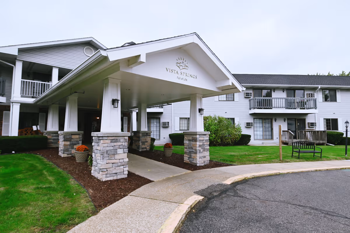Exterior view of a senior living facility with a covered entrance supported by stone pillars. The building is two stories with balconies and windows, surrounded by green grass and shrubs. There is a bench and a lamp post near the entrance driveway.