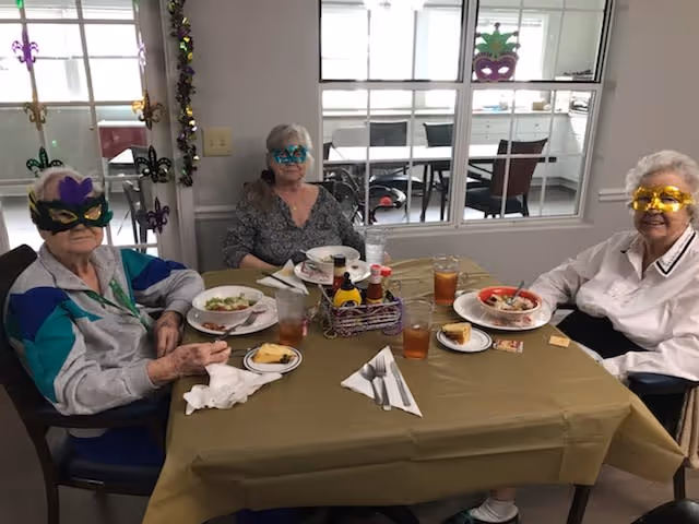 Three elderly women wearing colorful Mardi Gras masks sitting around a table with plates of food and drinks in a dining area decorated with Mardi Gras-themed ornaments on the window and wall.