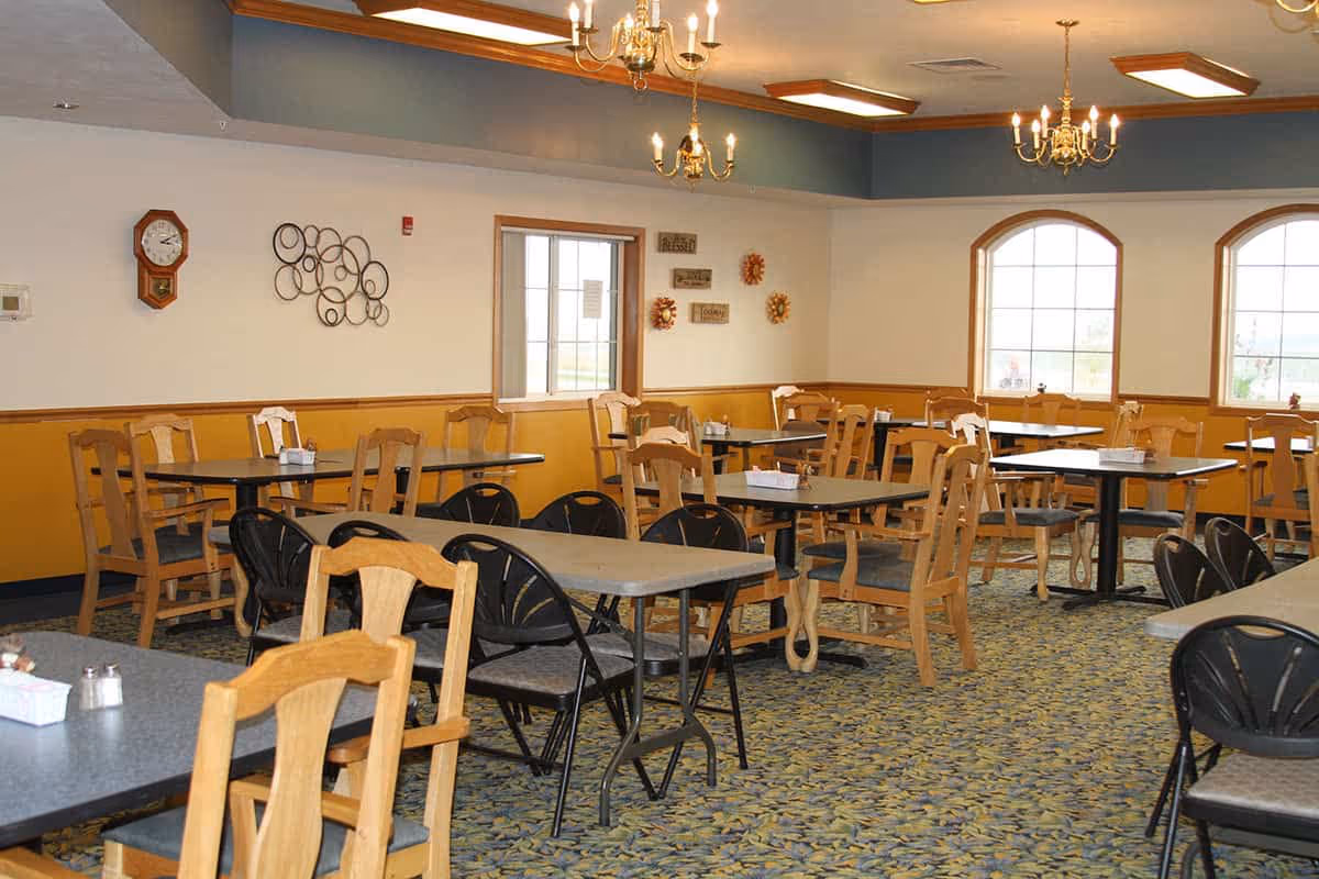 A dining room with multiple tables and chairs arranged neatly. The room features wooden chairs and black folding chairs around gray tables. The walls are painted white with a yellow lower half and decorated with wall art and a clock. There are large windows letting in natural light and chandeliers hanging from the ceiling.
