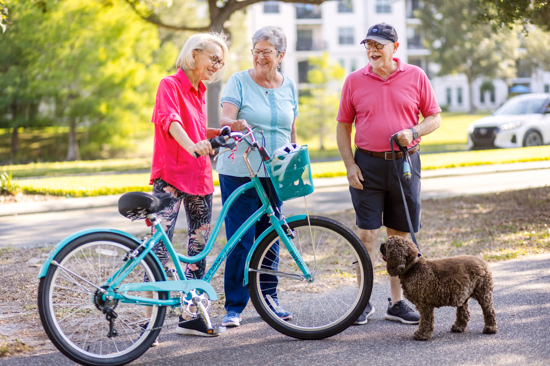 Three elderly people outdoors on a sunny day. Two women are standing next to a teal bicycle, smiling and talking. A man wearing a red shirt, black shorts, and a black cap is holding a leash attached to a small brown dog. Trees and a building are visible in the background.