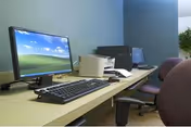 A computer workstation area with two desktop computers, keyboards, and mice on a long beige desk. There are office chairs in front of the desks and a small plant in the background against a blue wall.