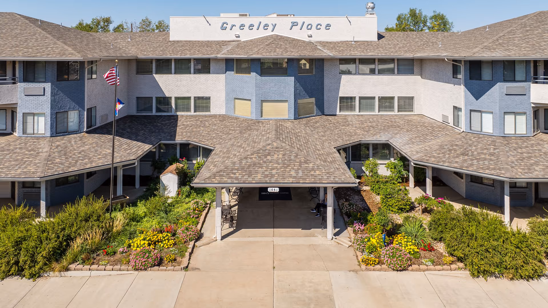 Front exterior view of a multi-story building named Greeley Place with a covered entrance, landscaped flower beds, and two flagpoles displaying the American and Colorado state flags.
