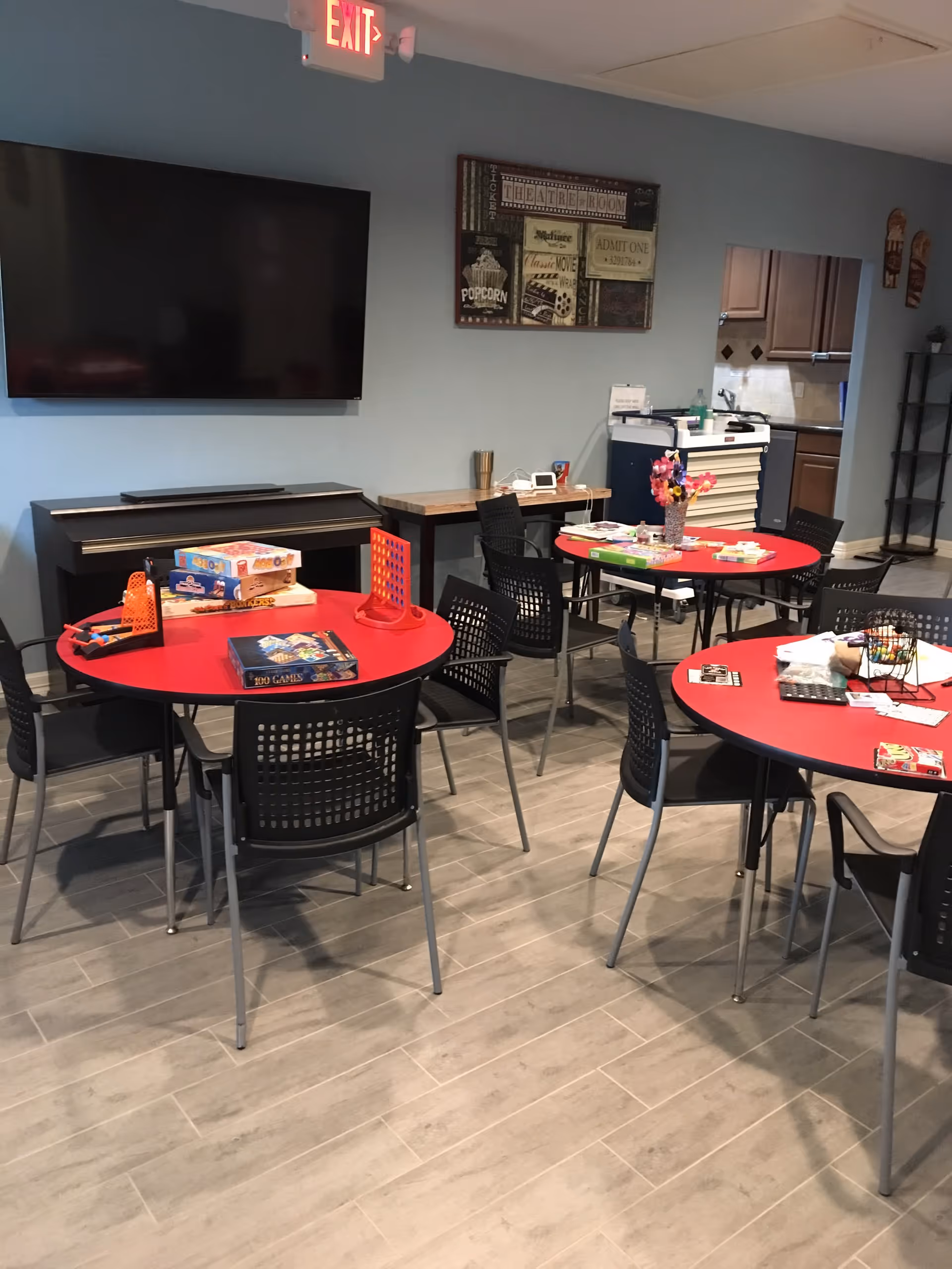 A common area with three round red tables surrounded by black chairs. Various board games and activity materials are placed on the tables. A large flat-screen TV is mounted on the wall above a piano or keyboard. A decorative sign with movie-themed artwork hangs on the wall. The floor is covered with light-colored tiles, and there is an exit sign above a door in the background.