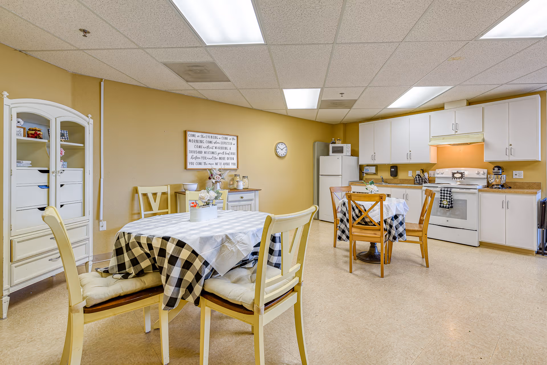 A bright kitchen and dining area in a senior living facility with two tables covered in checkered tablecloths and surrounded by chairs. The kitchen features white cabinets, a refrigerator, microwave, stove, and various kitchen appliances. A white cabinet with glass doors is on the left, and a wall clock and decorative sign are mounted on the beige wall.