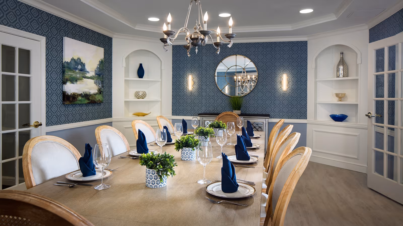A formal dining room with a long wooden table set for twelve people. Each place setting has a white plate, silverware, a wine glass, and a blue folded napkin. The room features blue patterned wallpaper, white built-in shelves with decorative items, a round mirror on the far wall, and a chandelier hanging above the table.