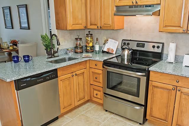 A modern kitchen with wooden cabinets, a stainless steel electric stove with a kettle on top, a dishwasher, a sink with a faucet, and granite countertops. There are decorative jars, a cookbook, and a paper towel holder on the counters. In the background, a dining area with a table and chairs is partially visible.