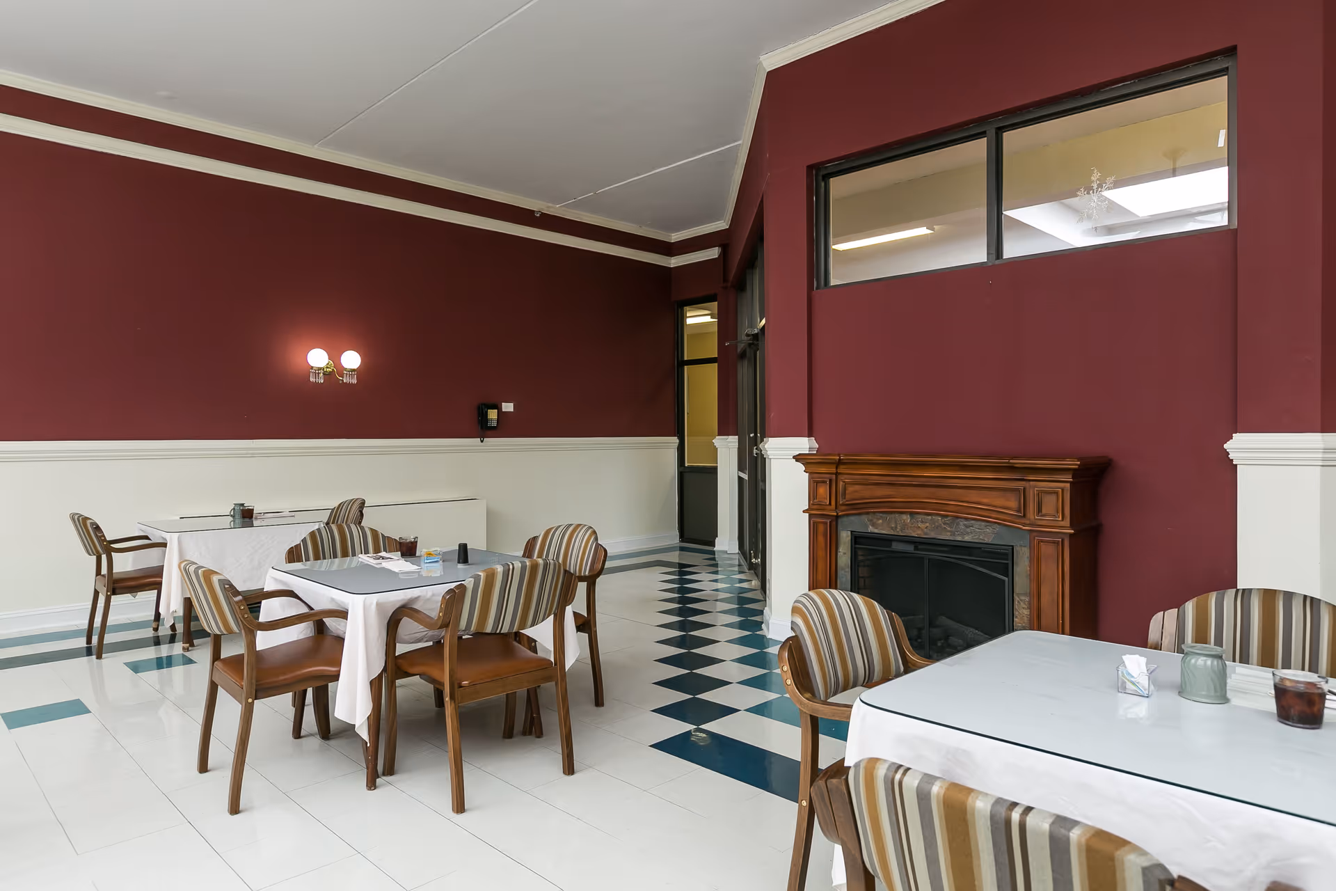 A dining area with tables covered in white tablecloths and striped cushioned chairs. The room has a maroon and white color scheme with a wooden fireplace on one wall and a checkered tile floor.