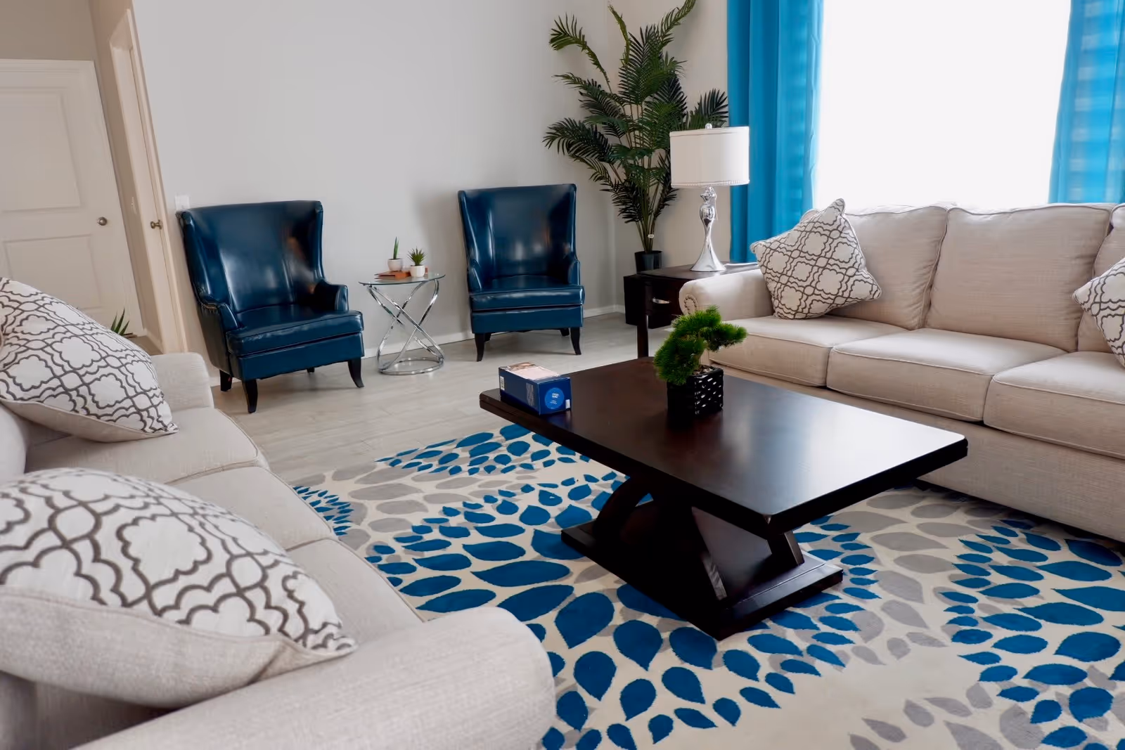 A bright living room with two beige sofas adorned with patterned pillows, two dark blue armchairs, a dark wooden coffee table with a small plant and a box on it, a side table with a lamp, a tall green plant in the corner, and blue curtains framing a window.
