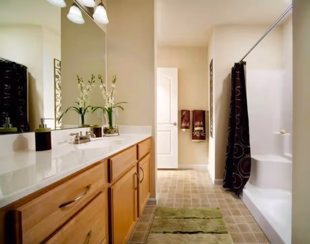 A clean and well-lit bathroom featuring a long countertop with a sink, wooden cabinets underneath, a large mirror above the sink, a vase with white flowers, a soap dispenser, a green bath mat on the tiled floor, a shower area with a dark shower curtain, and two decorative towels hanging on the wall near a closed door.