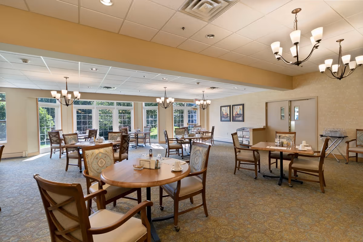 A bright and spacious dining room with multiple wooden tables and cushioned chairs arranged neatly. Large windows along one wall allow natural light to fill the room, showcasing a view of greenery outside. The ceiling has recessed lighting and several elegant chandeliers. The walls are decorated with framed artwork, and there is a small service cart with glasses and cups in the corner.