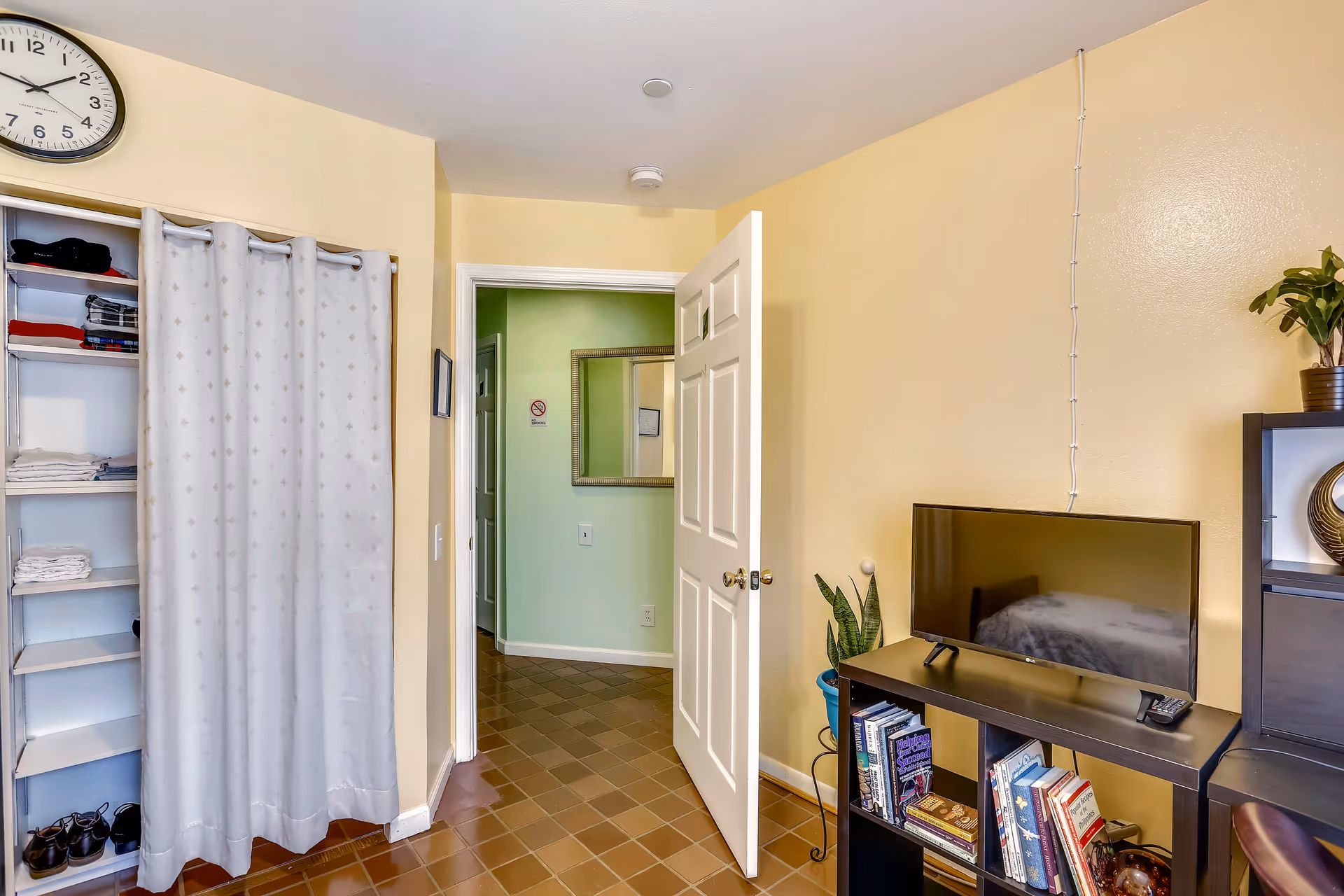 Interior view of a room with beige walls and brown tiled floor. There is an open white door leading to a hallway with light green walls. On the left side, there is a closet with shelves holding folded clothes and shoes, partially covered by a white curtain with a subtle pattern. On the right side, there is a black TV stand with a flat-screen TV on top, a remote control, and several books on the shelves below. A potted plant is placed next to the TV stand, and another plant is on a nearby black shelving unit.