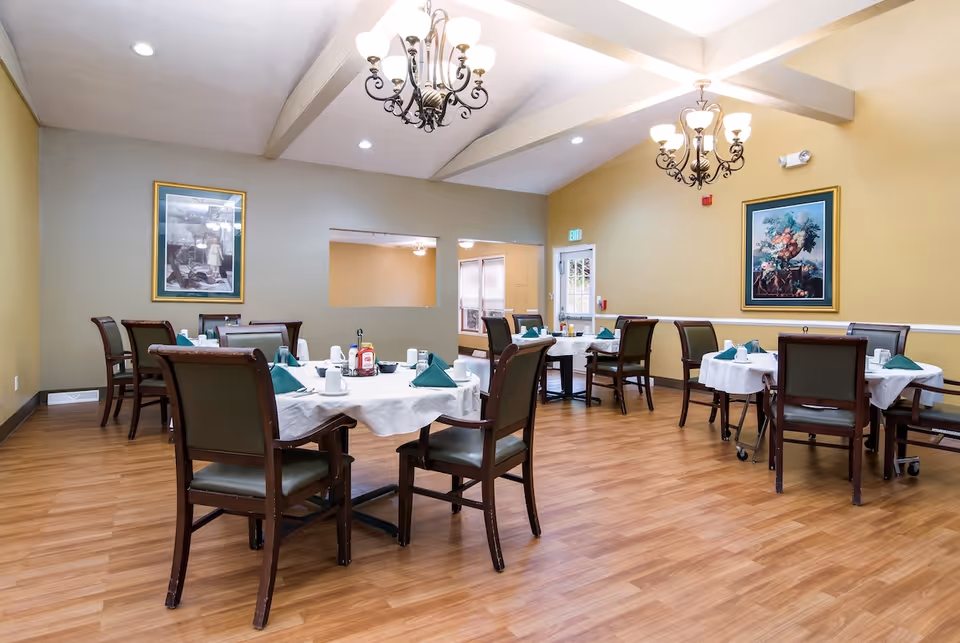 Dining room with round and rectangular tables set with white tablecloths and green napkins, wooden chairs, chandeliers, and framed artwork on the walls.