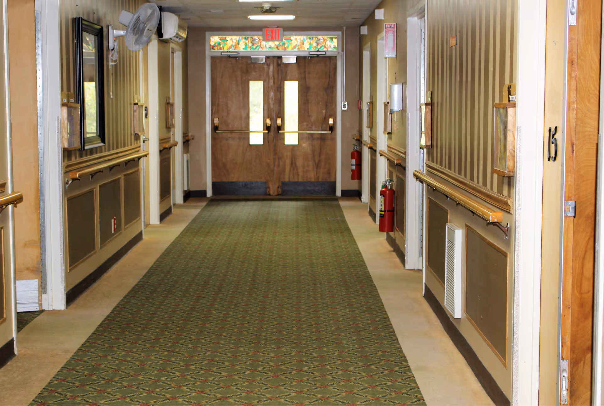 A long indoor hallway with patterned green carpet, beige walls with wooden handrails on both sides, several doors along the corridor, and double wooden exit doors with small windows at the end. There are fire extinguishers mounted on the walls and an exit sign above the doors.