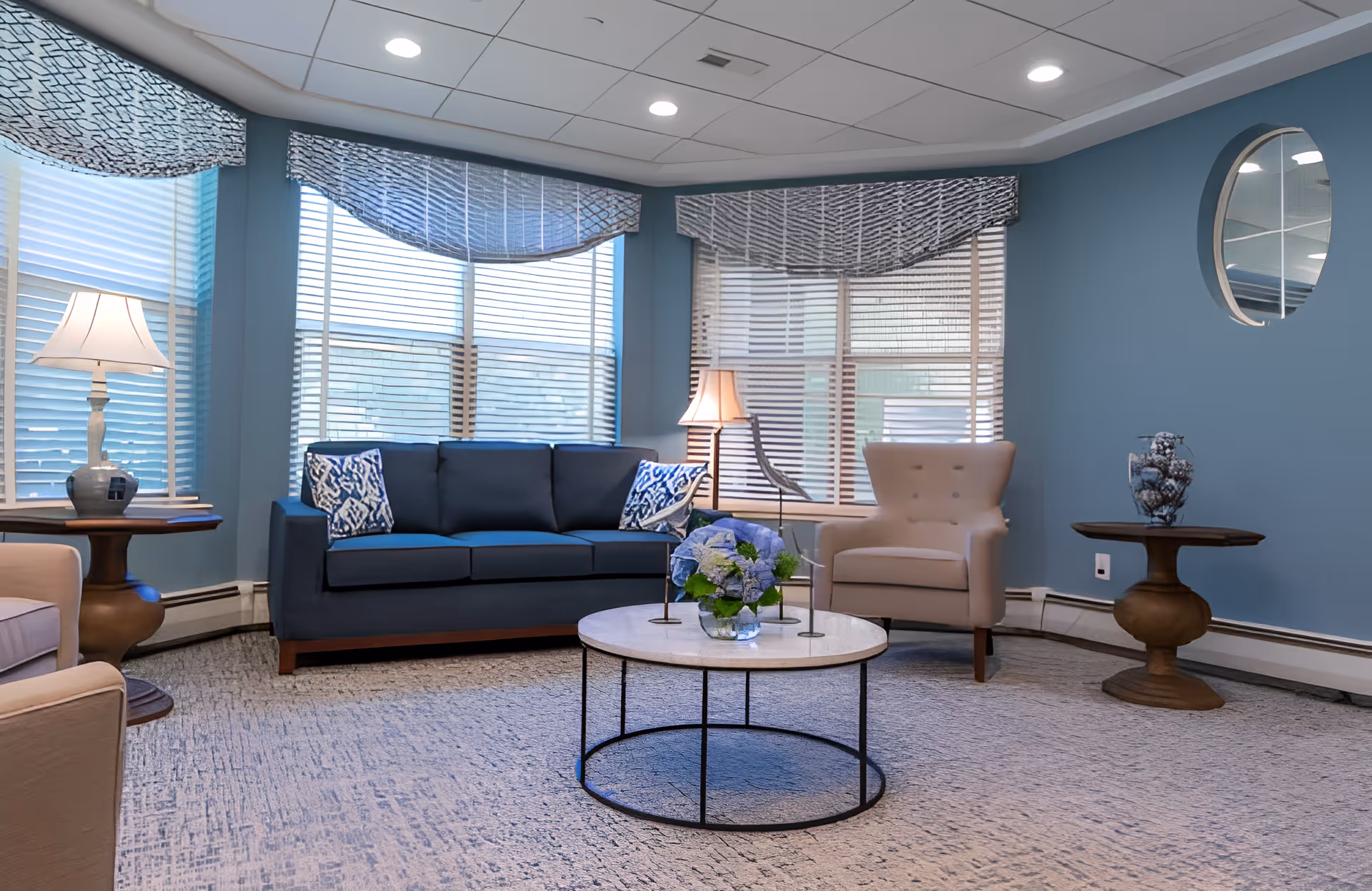 A cozy living room with light blue walls and a large window with blinds and patterned valances. The room features a dark blue sofa with patterned pillows, a beige armchair, two wooden side tables with lamps and decorative items, and a round coffee table with a floral arrangement. The ceiling has recessed lighting and a circular mirror is mounted on the wall.