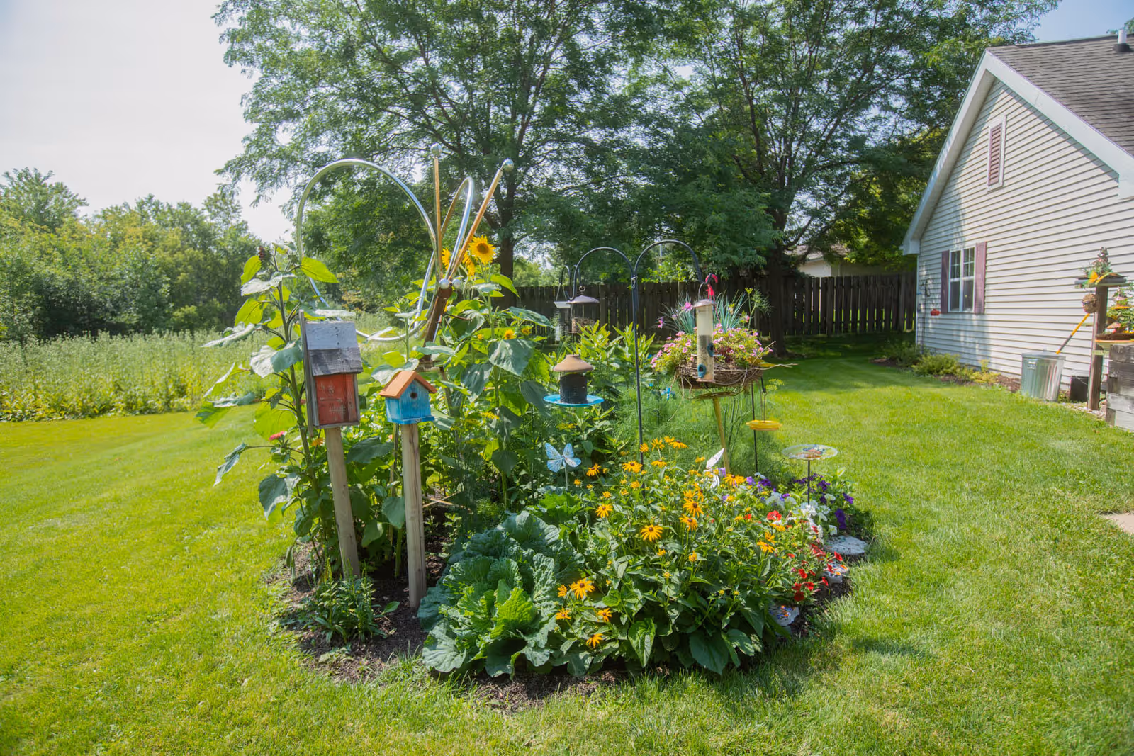 A vibrant garden area with various flowers, birdhouses, and bird feeders surrounded by green grass and trees, next to a light-colored house with purple shutters.