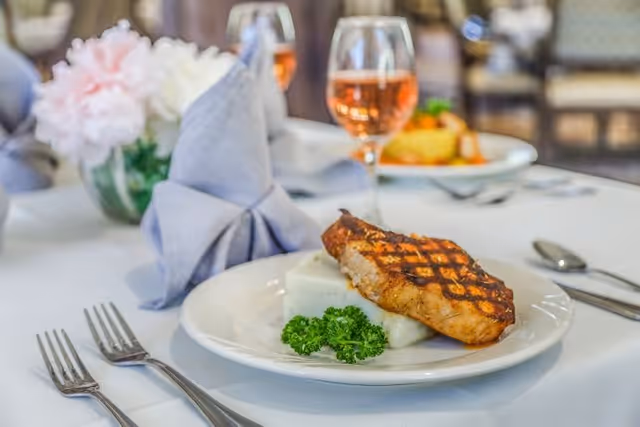 A close-up of a dining table set with a plate of grilled salmon, mashed potatoes, and a garnish of parsley. There is a glass of rosé wine and a folded gray cloth napkin on the table, with another plate of food and floral centerpiece blurred in the background.