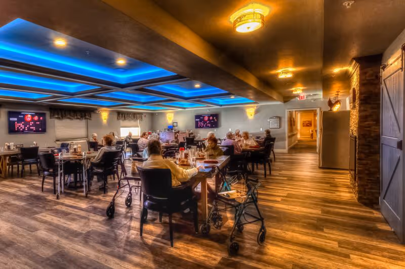 A dining room in a senior living facility with several elderly residents seated at tables. The room features wooden flooring, modern ceiling lights with blue accent lighting, and wall-mounted televisions. Walkers are visible next to some chairs.