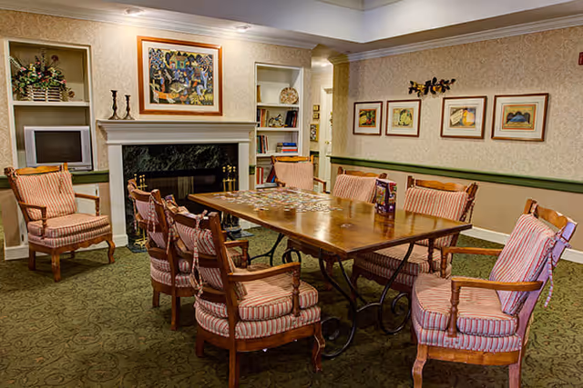 A cozy common room with a long wooden table surrounded by eight cushioned wooden chairs with striped upholstery. The room features a fireplace with a dark marble surround, built-in shelves with decorative items, framed artwork on the walls, and a green patterned carpet.