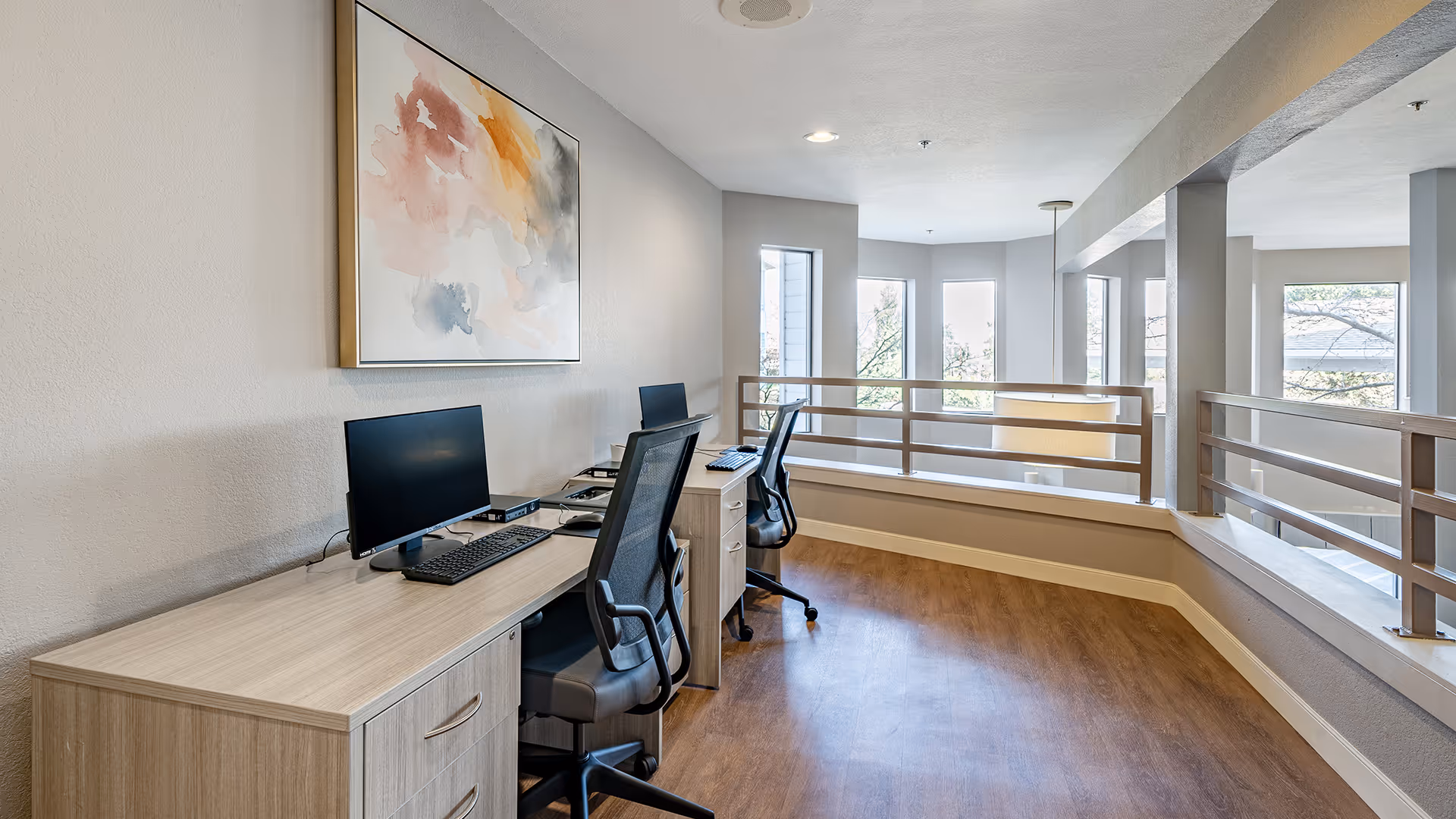 A bright office nook with two desks, each equipped with a computer monitor, keyboard, and office chair. The space has light-colored walls, wood flooring, and large windows letting in natural light. A colorful abstract painting hangs on the wall above one desk. The area overlooks a lower level through a railing.