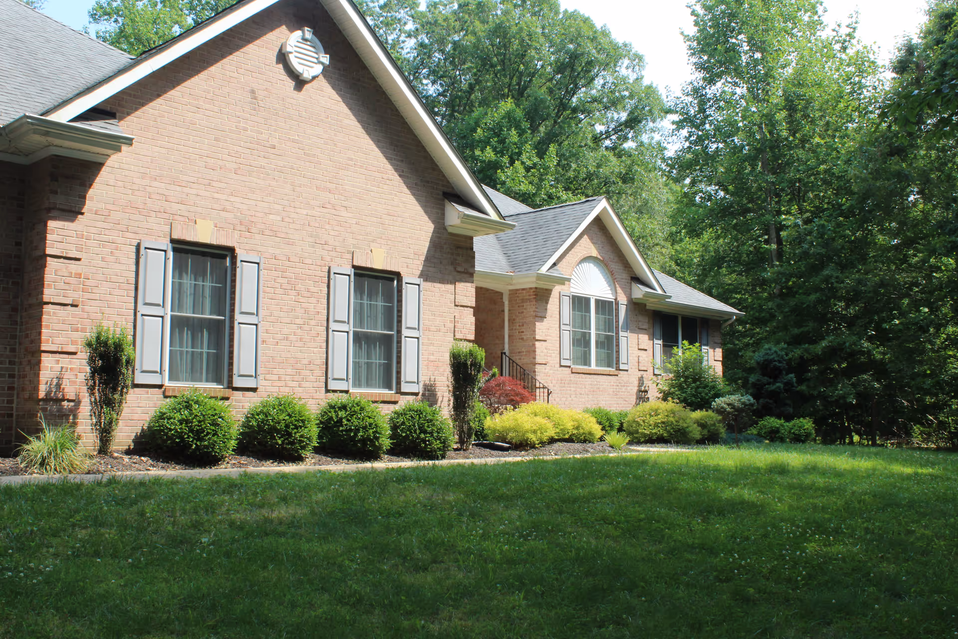 Exterior view of a single-story brick building with multiple windows, surrounded by green bushes and trees, with a well-maintained grassy lawn in front.