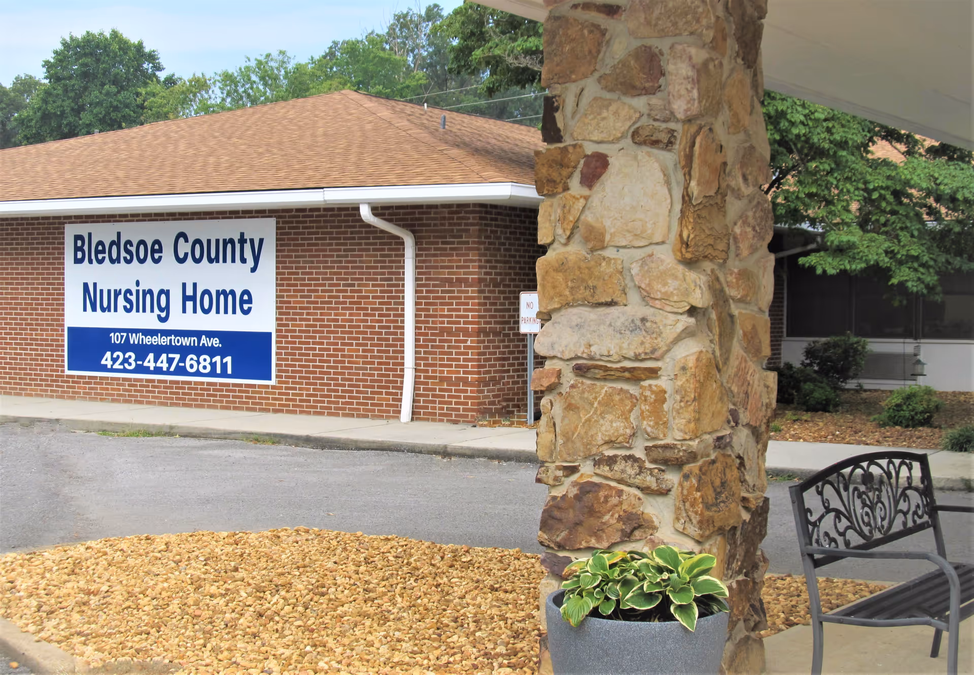 Exterior view of Bledsoe County Nursing Home showing a brick building with a large sign displaying the facility's name, address, and phone number. In the foreground, there is a stone pillar, a potted plant, and a metal bench under a covered area.