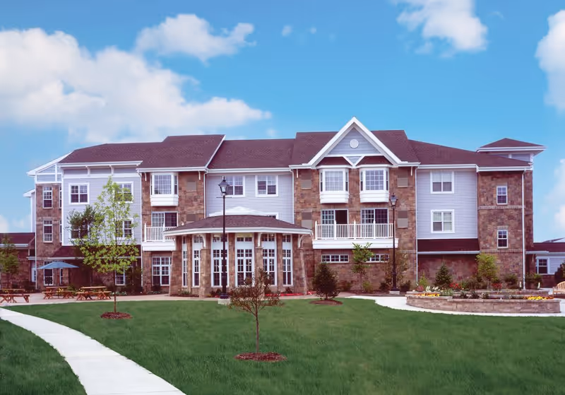Front exterior view of a multi-story retirement community building with stone and siding facade, large windows, balconies, and a well-maintained lawn with young trees and a curved sidewalk under a blue sky with scattered clouds.