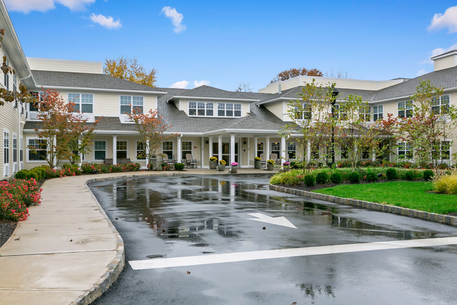 Exterior view of a senior living facility with a wet driveway, landscaped garden, and a two-story building featuring multiple windows and a covered entrance with columns. The sky is blue with some clouds.