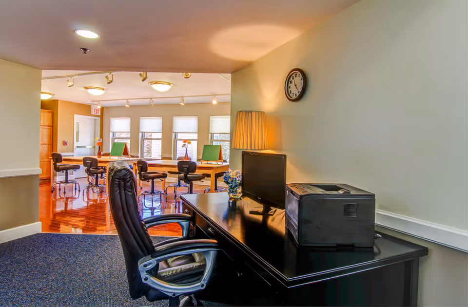 Interior common area with a desk, office chair, printer and lamp in the foreground and a communal table with stools and windows in the background.