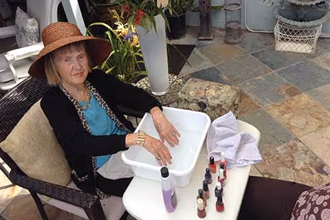 An older woman wearing a sun hat sits in a wicker chair on a tiled patio with her hands soaking in a white basin surrounded by nail polish bottles, a towel, and potted plants.