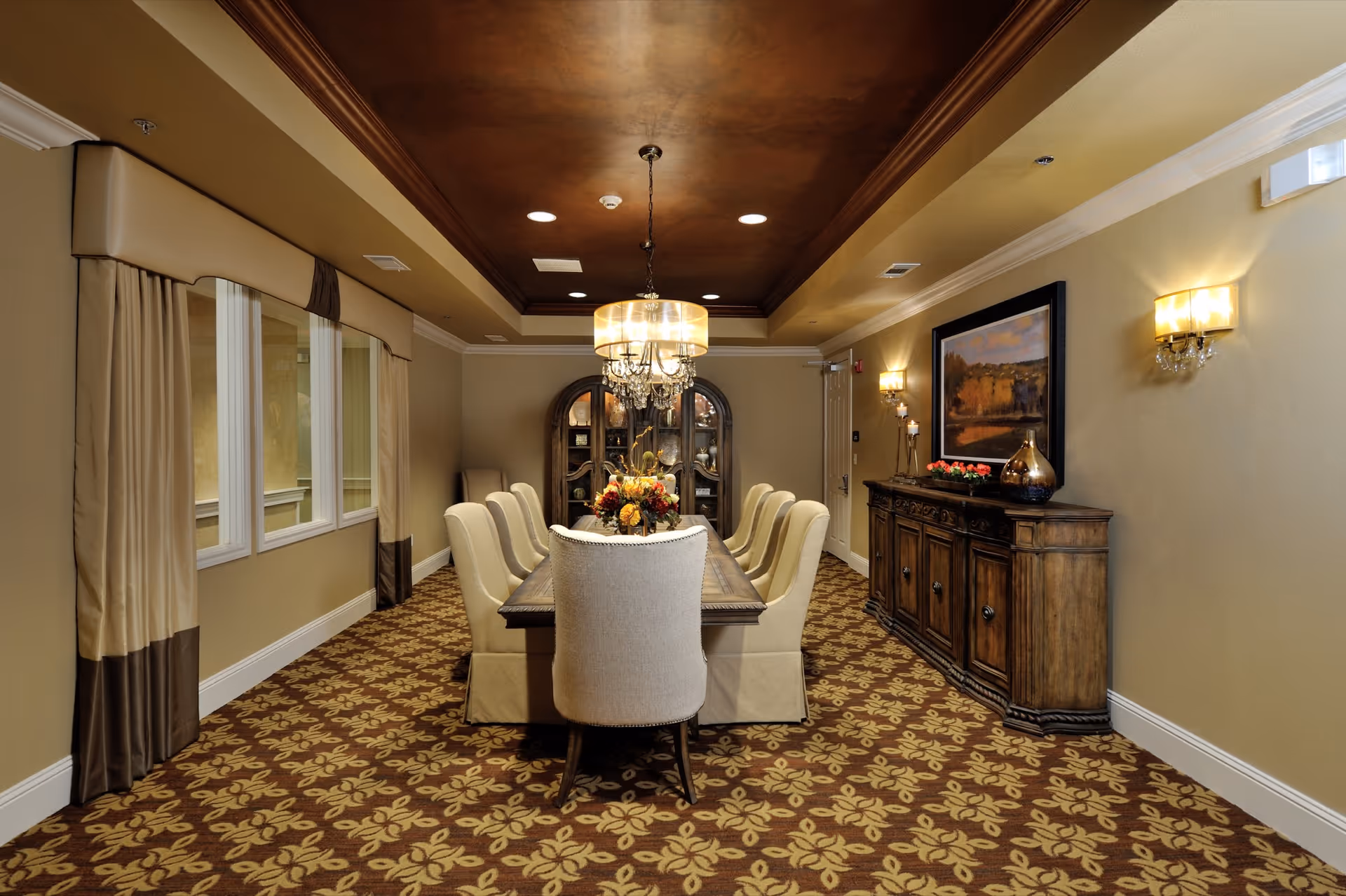 Formal dining room with a long table surrounded by upholstered chairs, a chandelier overhead, patterned carpet, and a wooden sideboard.