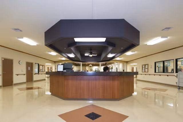 Interior view of a reception desk area in a healthcare facility with a large wooden counter, overhead lighting, and beige walls with doors and windows along the hallway.