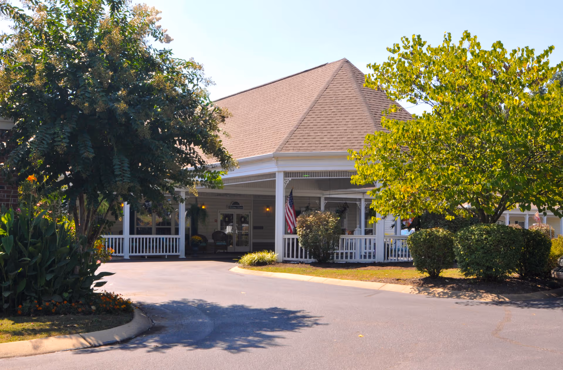 Exterior view of Charter Senior Living of Jackson showing the entrance with a covered porch, white railings, and an American flag. The building is surrounded by trees and bushes with a paved driveway in front.