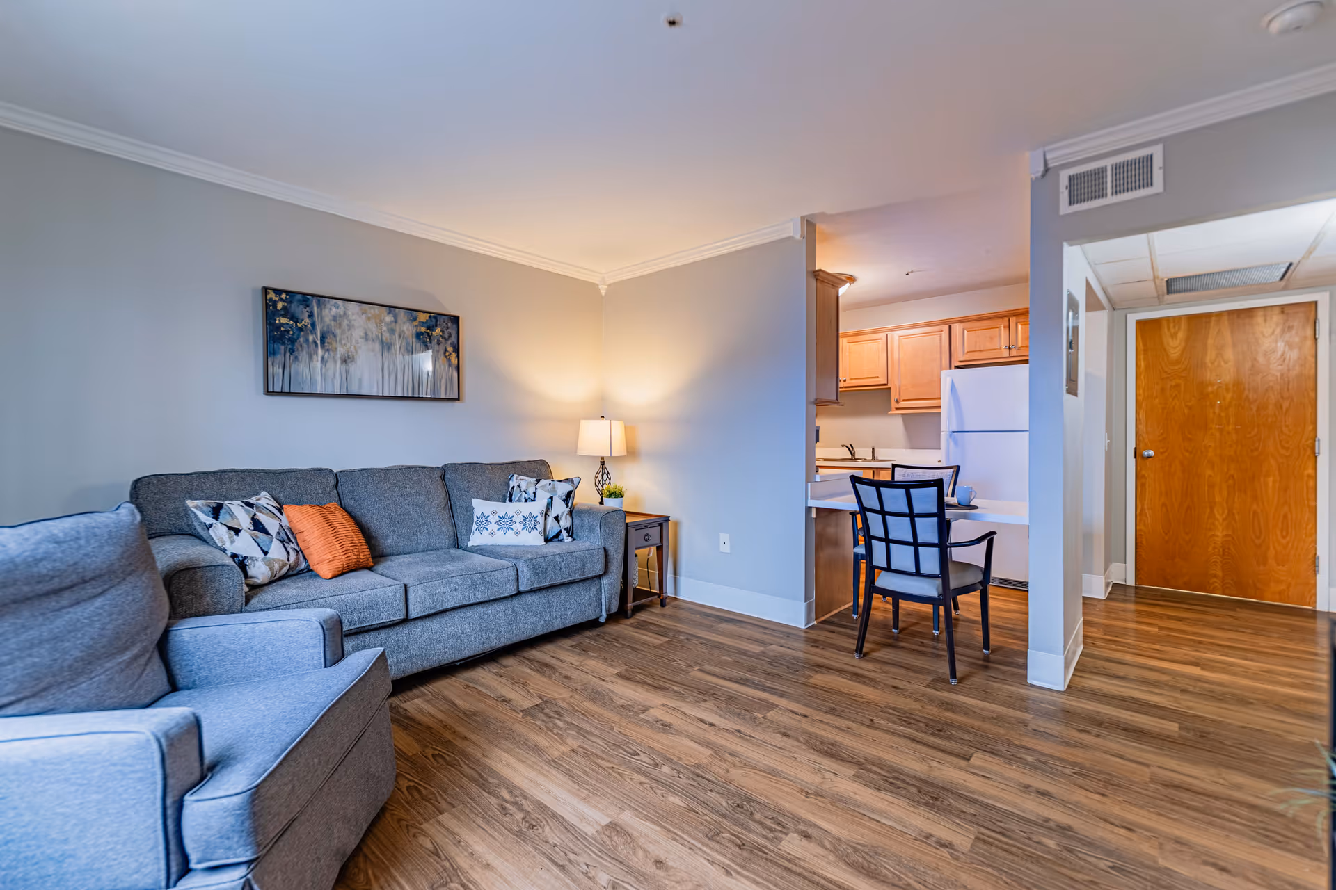 Cozy living room with a gray sofa and armchair, a side table with lamp, and a small kitchen and dining area visible in the background.