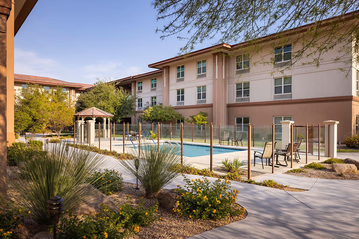 Outdoor area of Belmont Village Senior Living Scottsdale featuring a fenced swimming pool surrounded by chairs and tables, landscaped garden with desert plants and flowers, and a three-story building in the background under a clear blue sky.