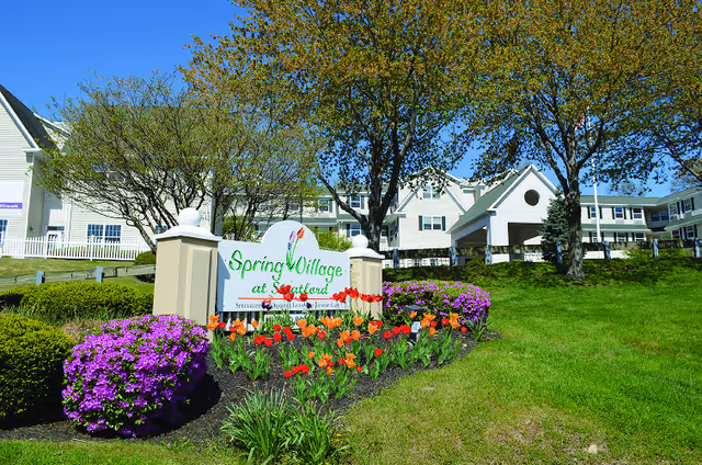 Exterior view of Spring Village Stratford senior living facility with a landscaped garden featuring colorful flowers and green grass in front of a large white building under a clear blue sky.