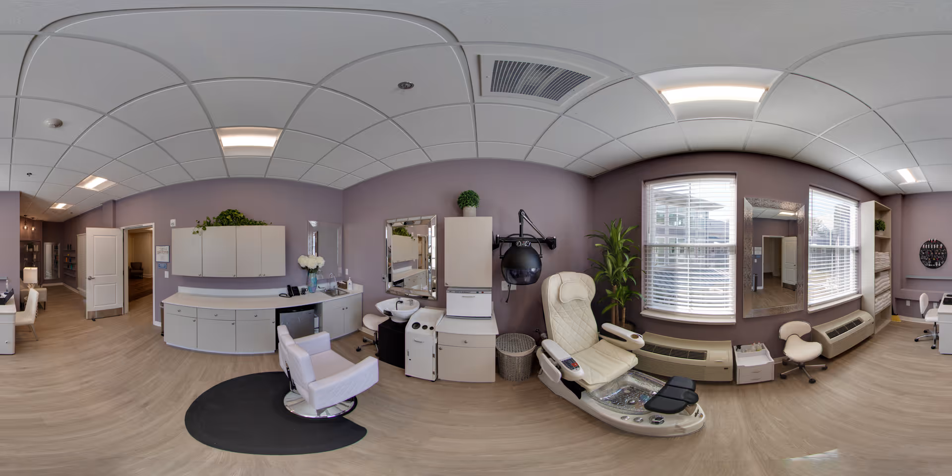 Interior view of a well-lit salon area in an assisted living facility with a pedicure chair, a salon chair, a hair dryer, cabinets, a sink, and large windows with blinds. The walls are painted light purple and the floor is light wood.